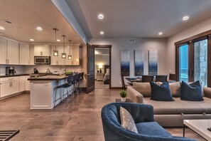 A modern kitchen with white cabinets, a central island, and pendant lighting. There is a dining area with a sofa and chairs, and a large window offering a view.
