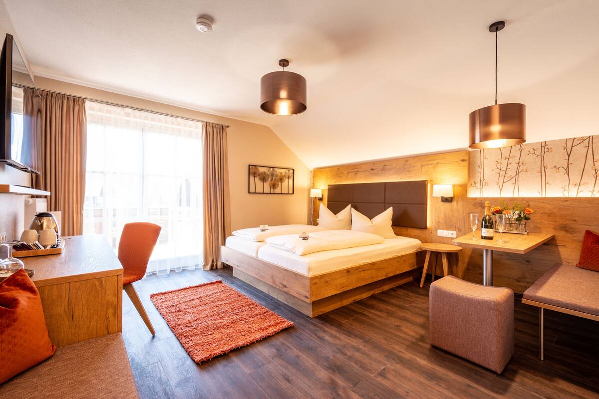 A modern hotel room with a wooden bed, a desk with a chair, a small table with a lamp, and a large window with curtains.