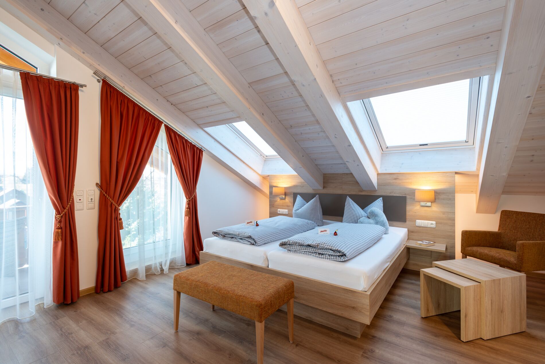 Un dormitorio tipo loft moderno con techo inclinado, vigas de madera, una cama grande con ropa de cama a cuadros, un banco, una pequeña mesa de madera y vistas de la ciudad a través de amplias ventanas.
