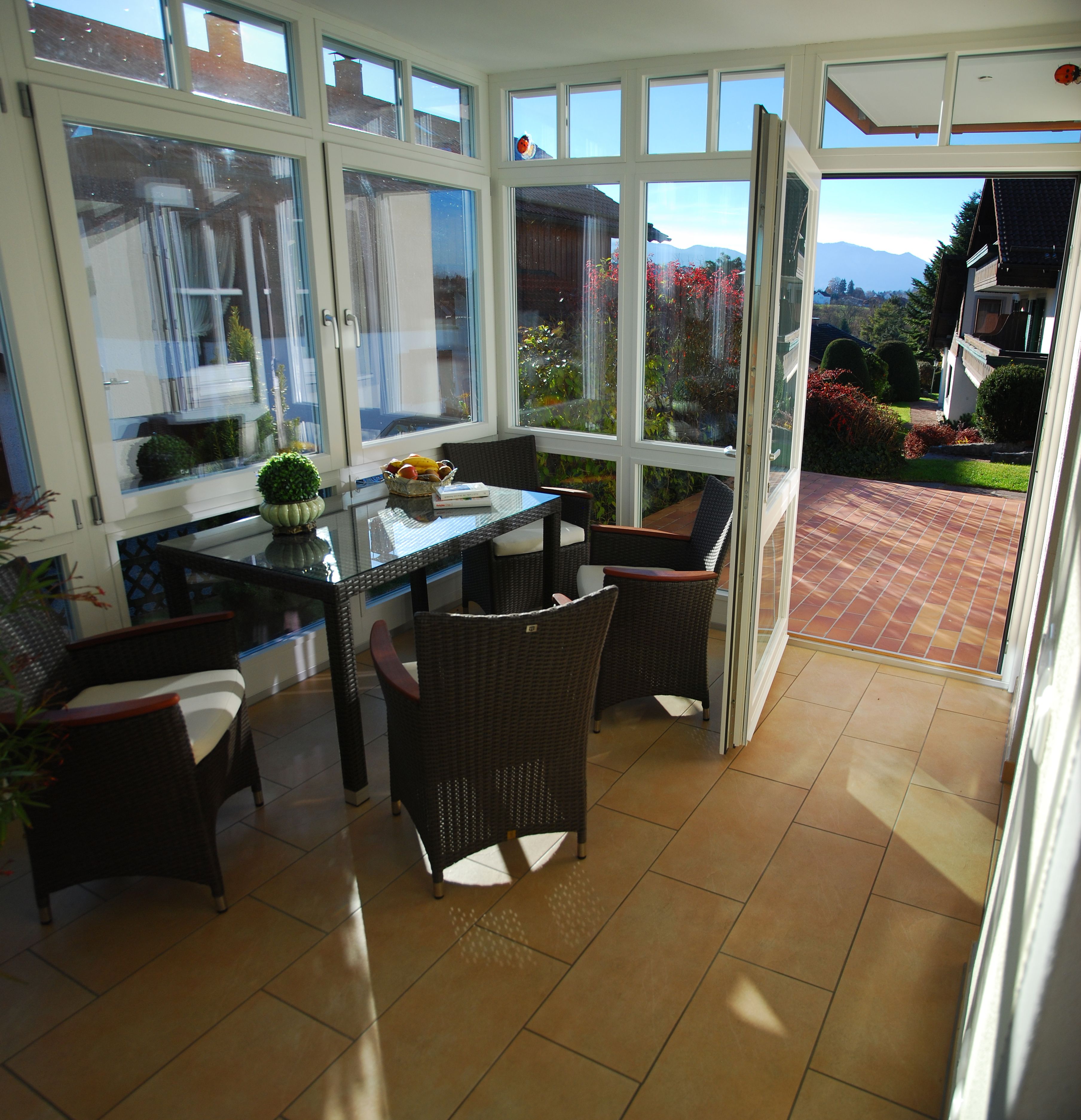 Eine sonnendurchflutete Terrasse mit Glastisch, Korbstühlen und Blick auf Garten und Berge.