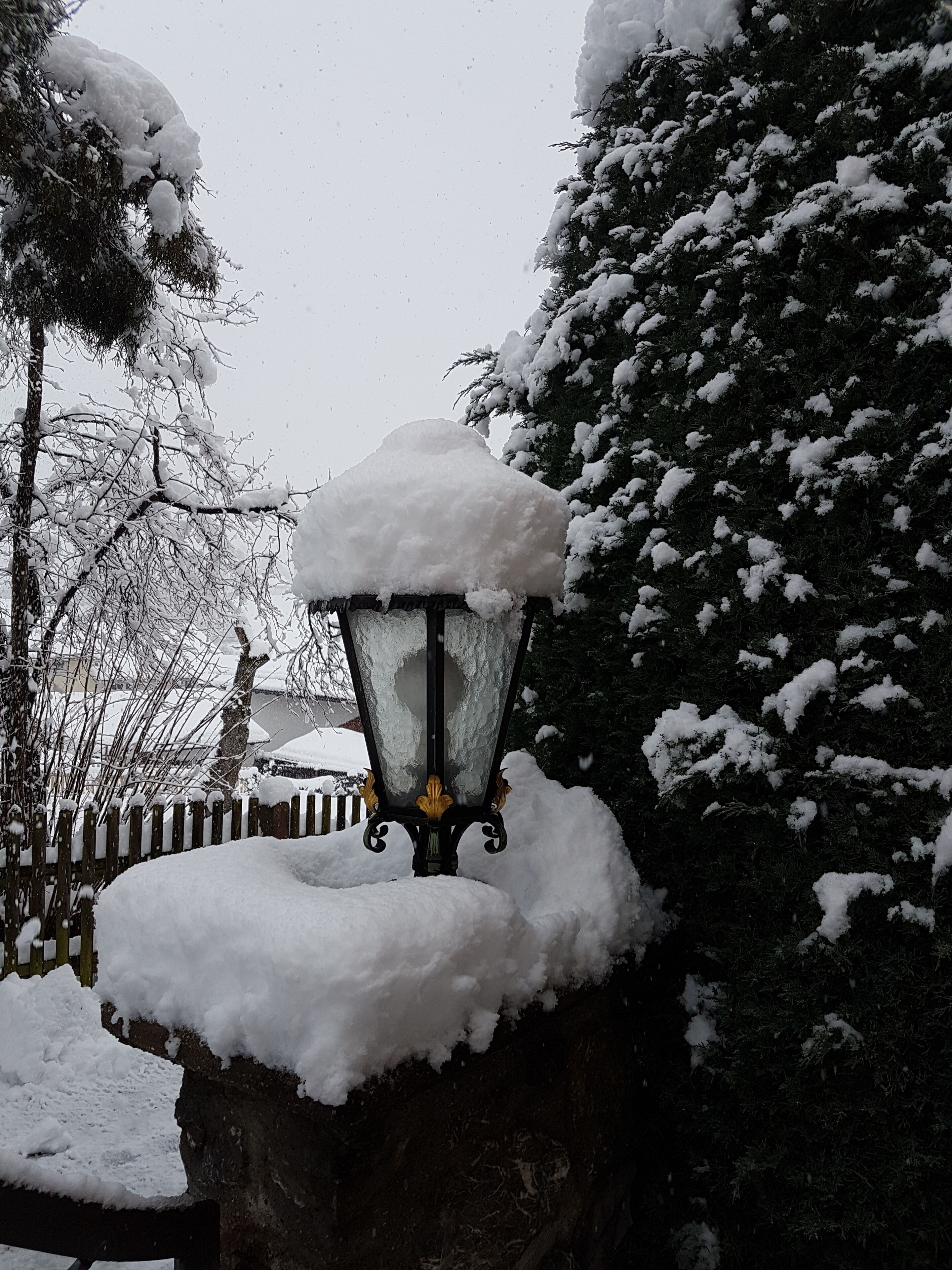 Un lampadaire enneigé, doté d’un socle en pierre, entouré d’arbres chargés de neige et d’une clôture en bois.