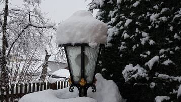 Un lampadaire enneigé, doté d’un socle en pierre, entouré d’arbres chargés de neige et d’une clôture en bois.