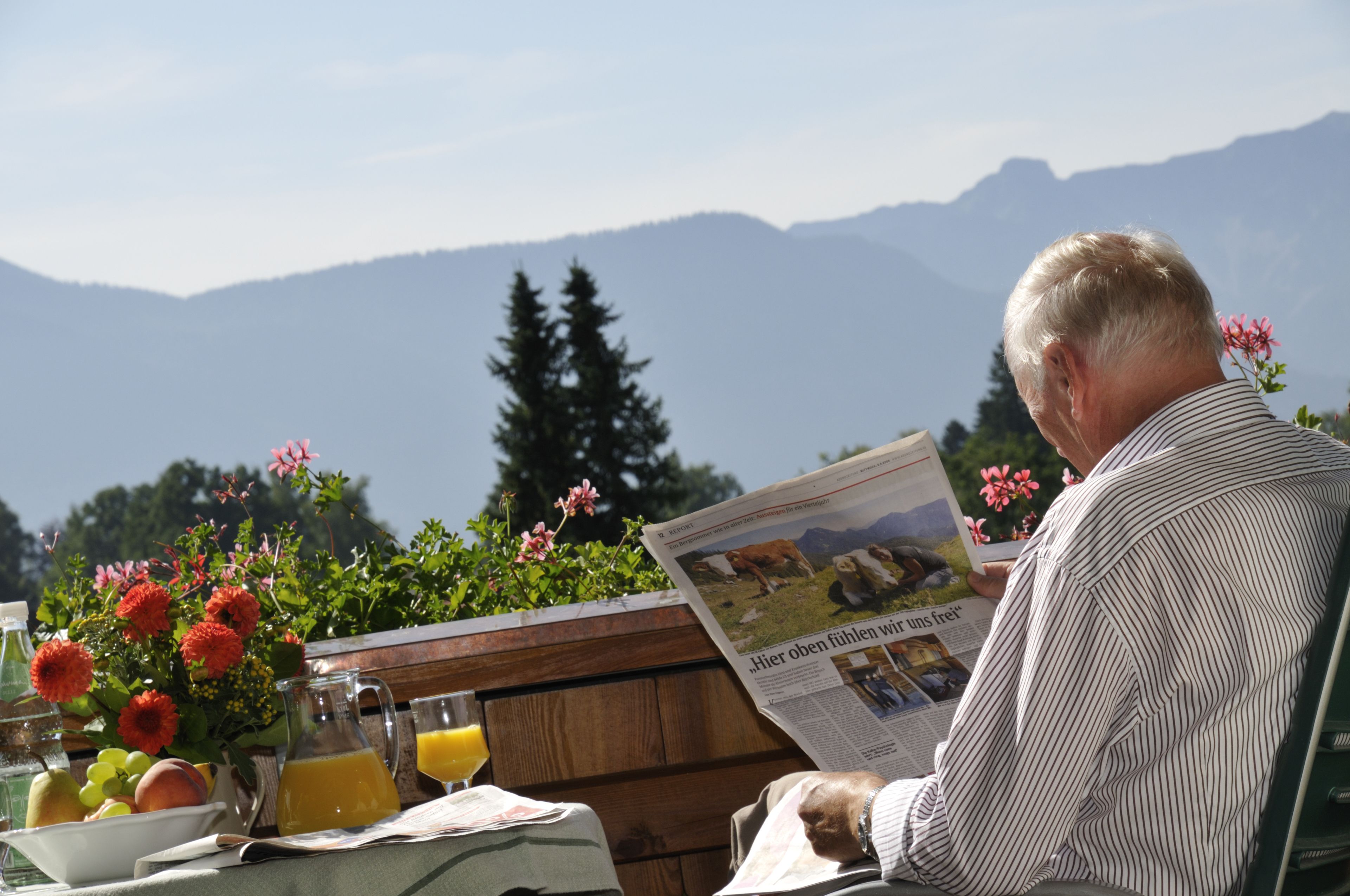 Un hombre leyendo un periódico en una mesa, con vistas a montañas al fondo.