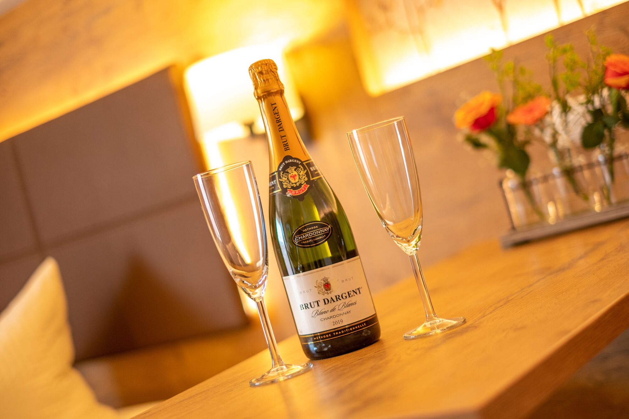 Uma garrafa de Brut Dargent Chardonnay 2019 sobre uma mesa de madeira com dois copos de vinho vazios.