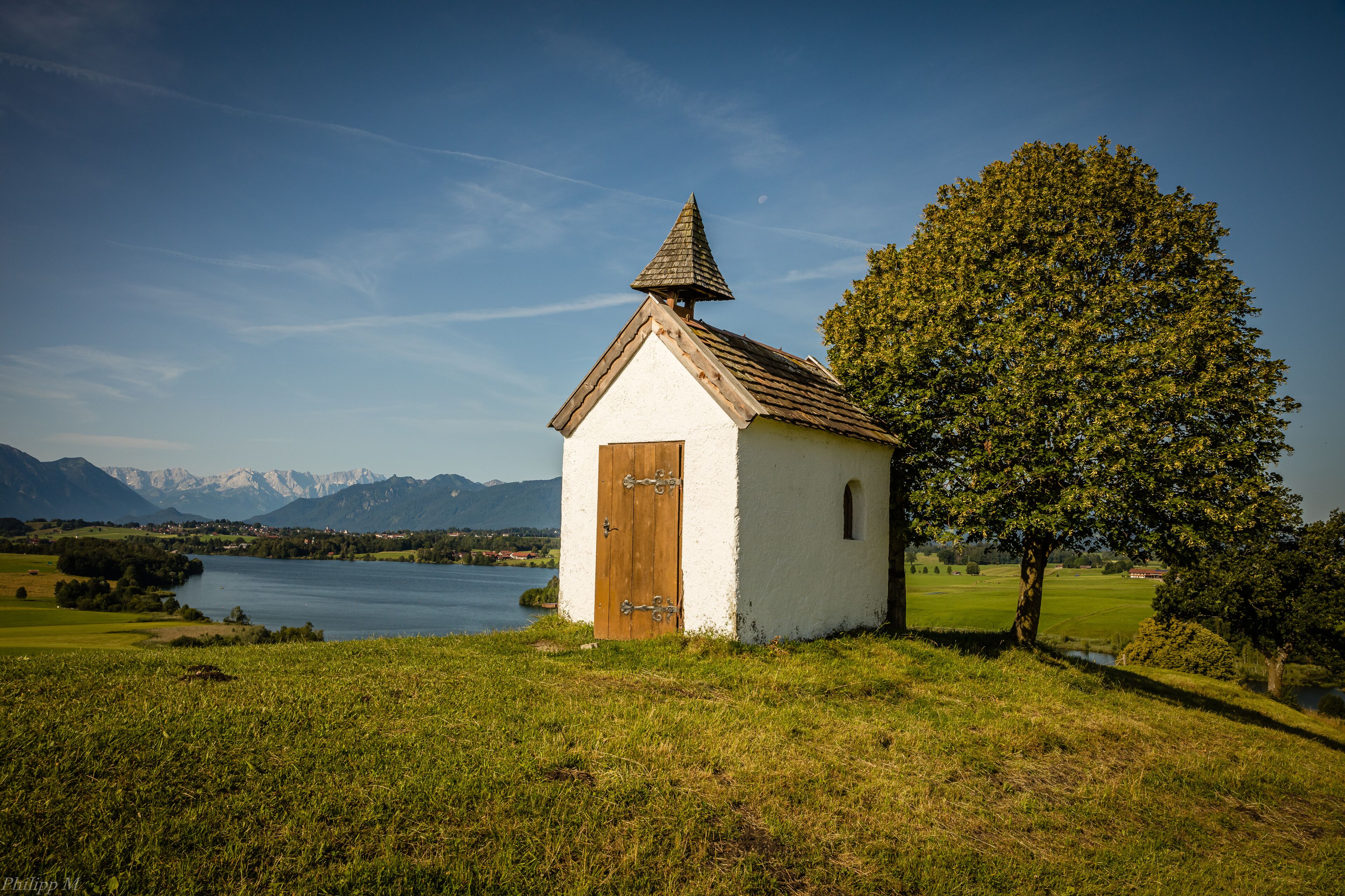 Eine kleine weiße Kirche mit Holztür und Kegeldach, umgeben von Gras und einem großen Baum, mit Bergen im Hintergrund.