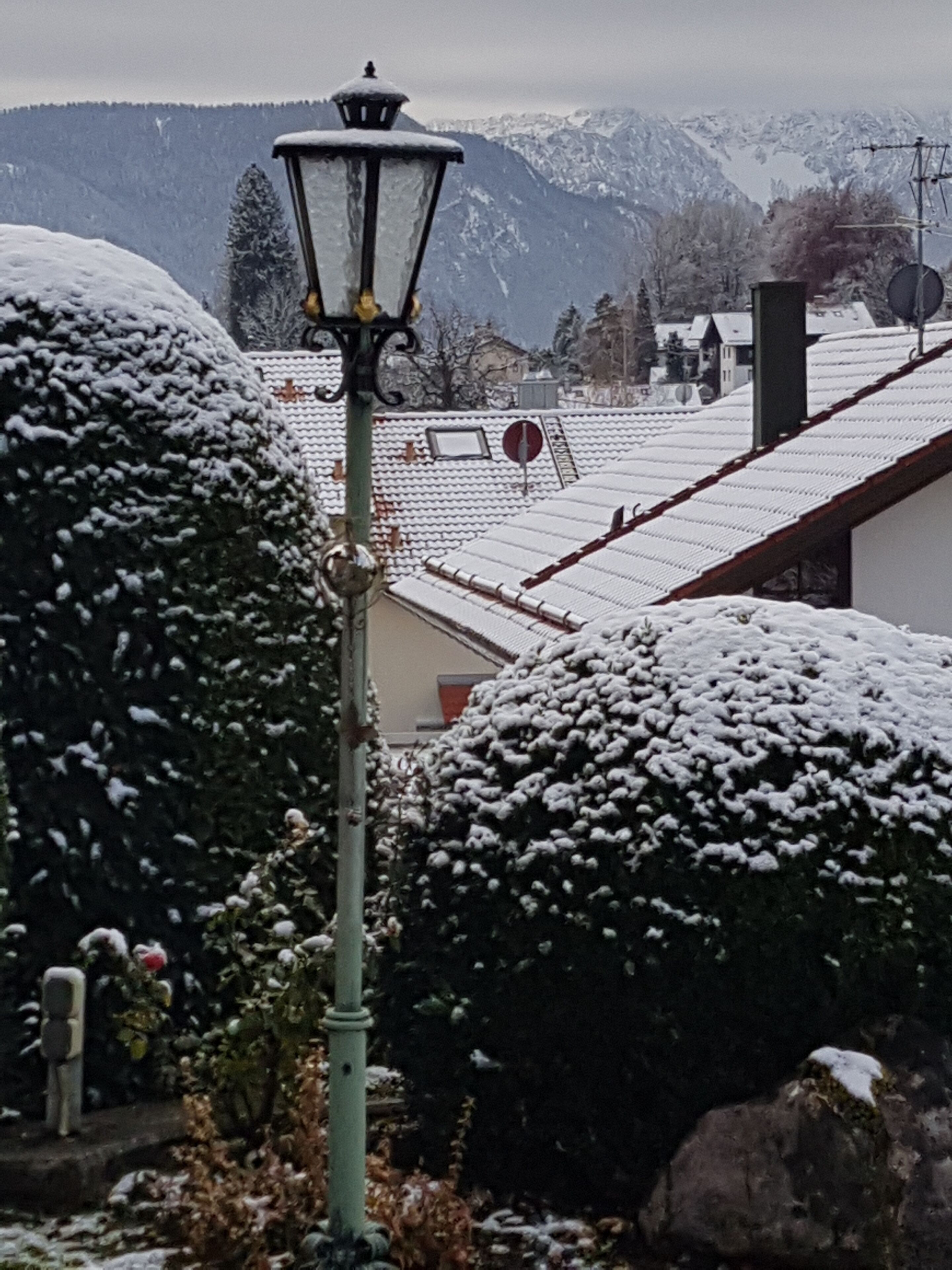 Lampione e case innevate con neve sui tetti, circondati da alberi imbiancati e montagne sullo sfondo.