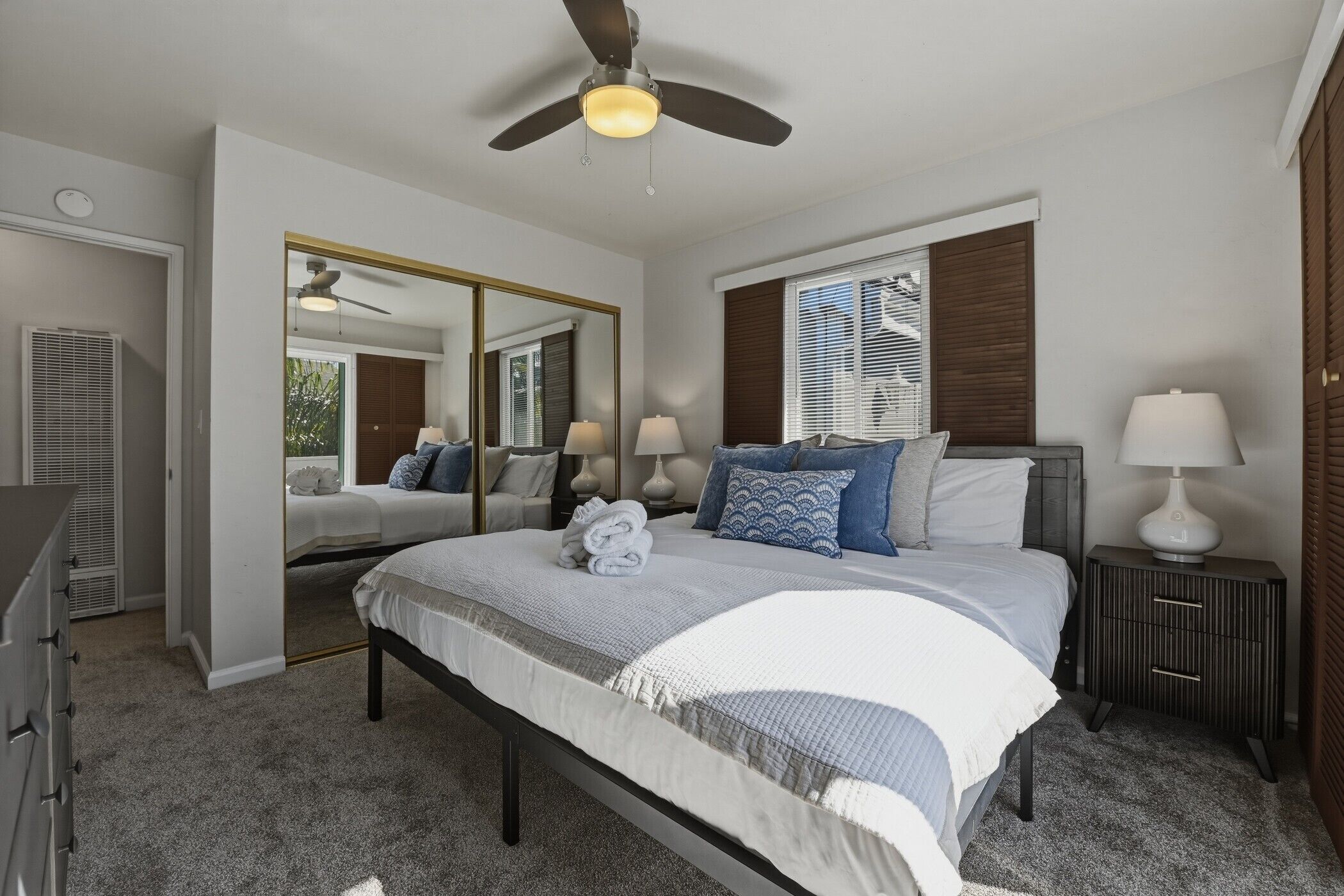 A bedroom with a bed, bedside lamps, a ceiling fan, a mirror, and a window with blinds.