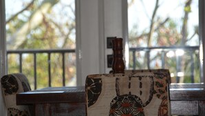 Un banco de madera con un cojín decorativo, una pequeña mesa de madera con tablero estampado, y vistas a árboles a través de una ventana con marco blanco.