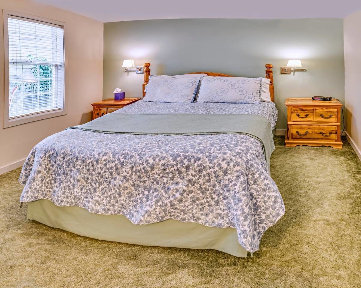 Une chambre avec un grand lit, deux tables de chevet, une commode, une fenêtre avec des stores et une lampe sur chaque table.