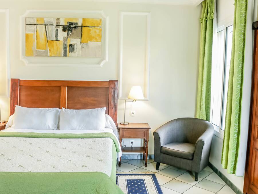 Una habitación de hotel con cama, una silla, una mesita de noche, una lámpara y una ventana con cortinas.