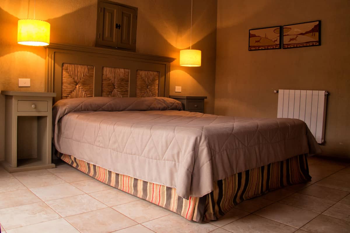 A bedroom with a bed, two bedside tables, a headboard, wall-mounted lamps, a radiator, and two wall art pieces.