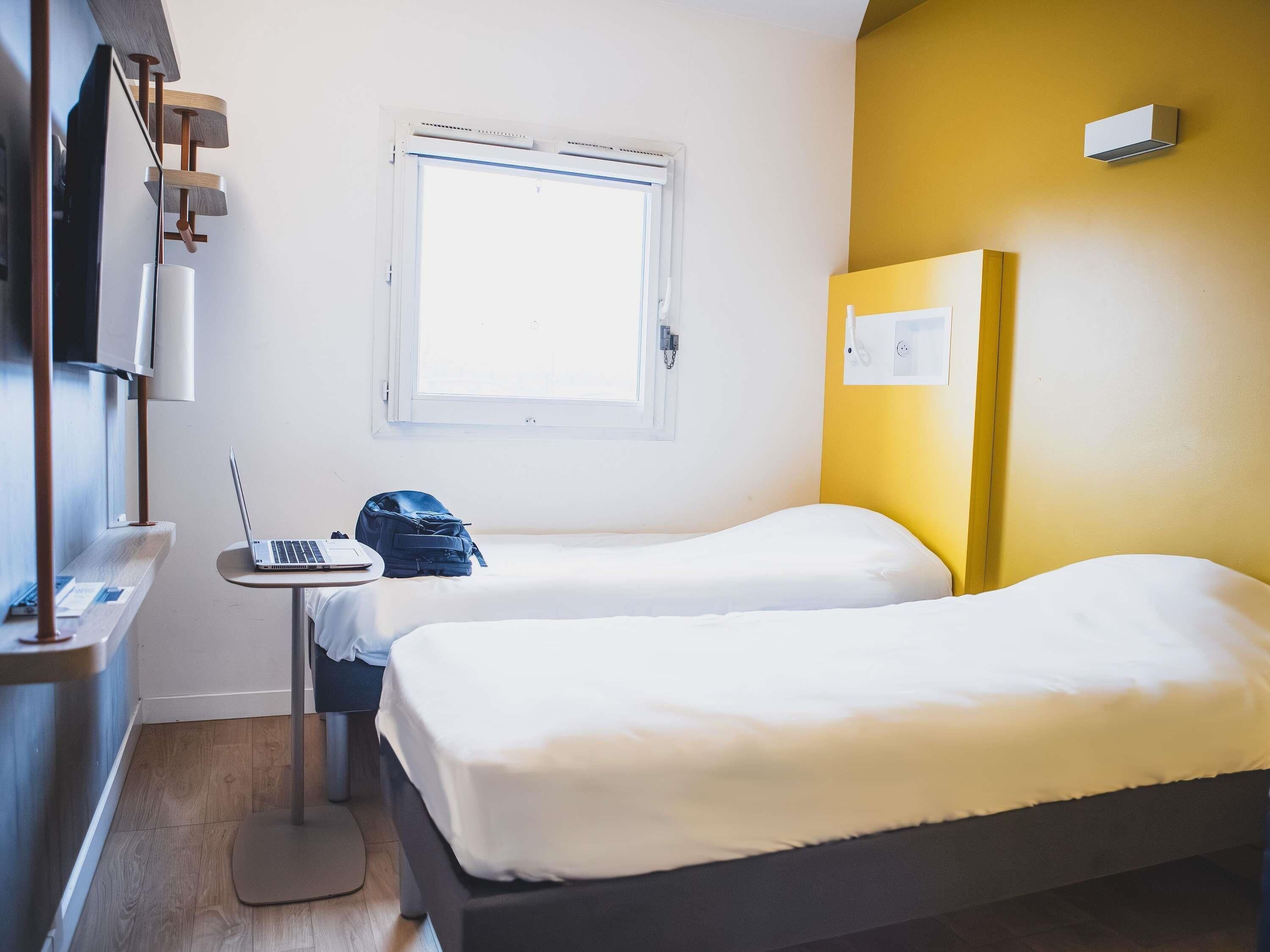 Et hotelværelse med to senge, et skrivebord med en bærbar computer, et vindue og en vægmonteret klimaanlæg.