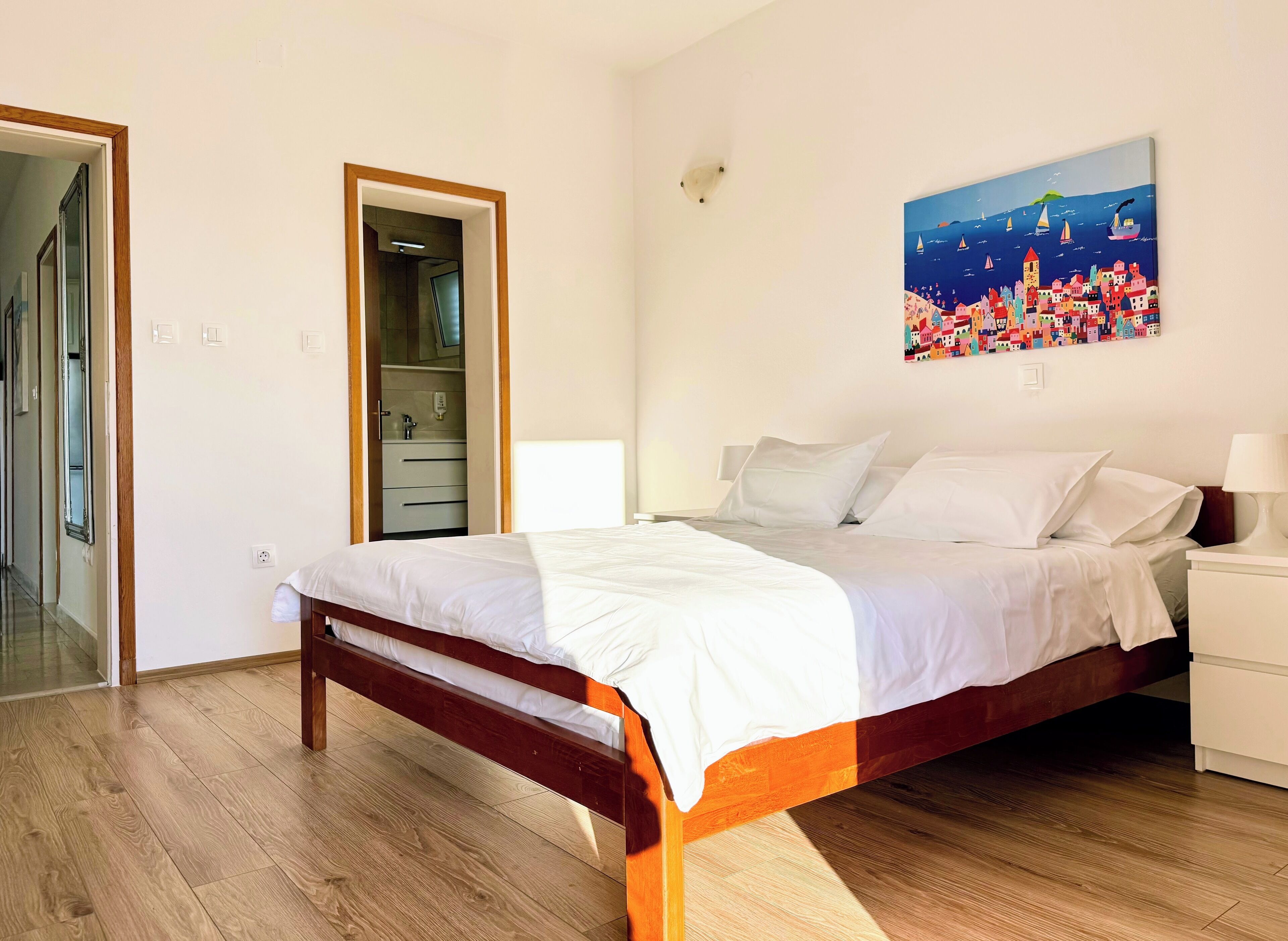 A bedroom with a wooden bed, white bedding, a nightstand, and a painting of a coastal town on the wall.