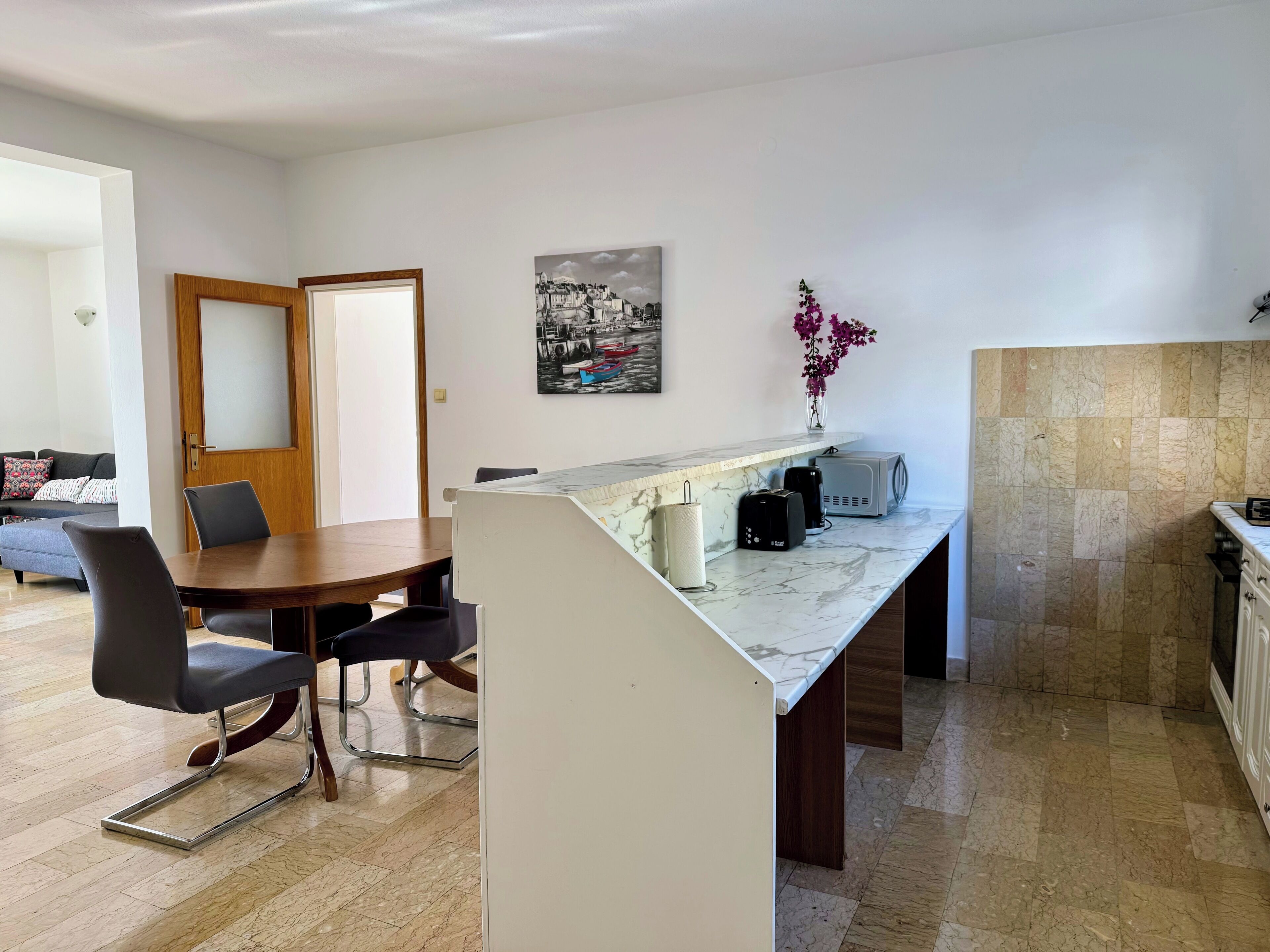 Een moderne keuken met een marmeren aanrecht, een eethoek met een houten tafel en stoelen, en een zithoek met een bank en een deur.