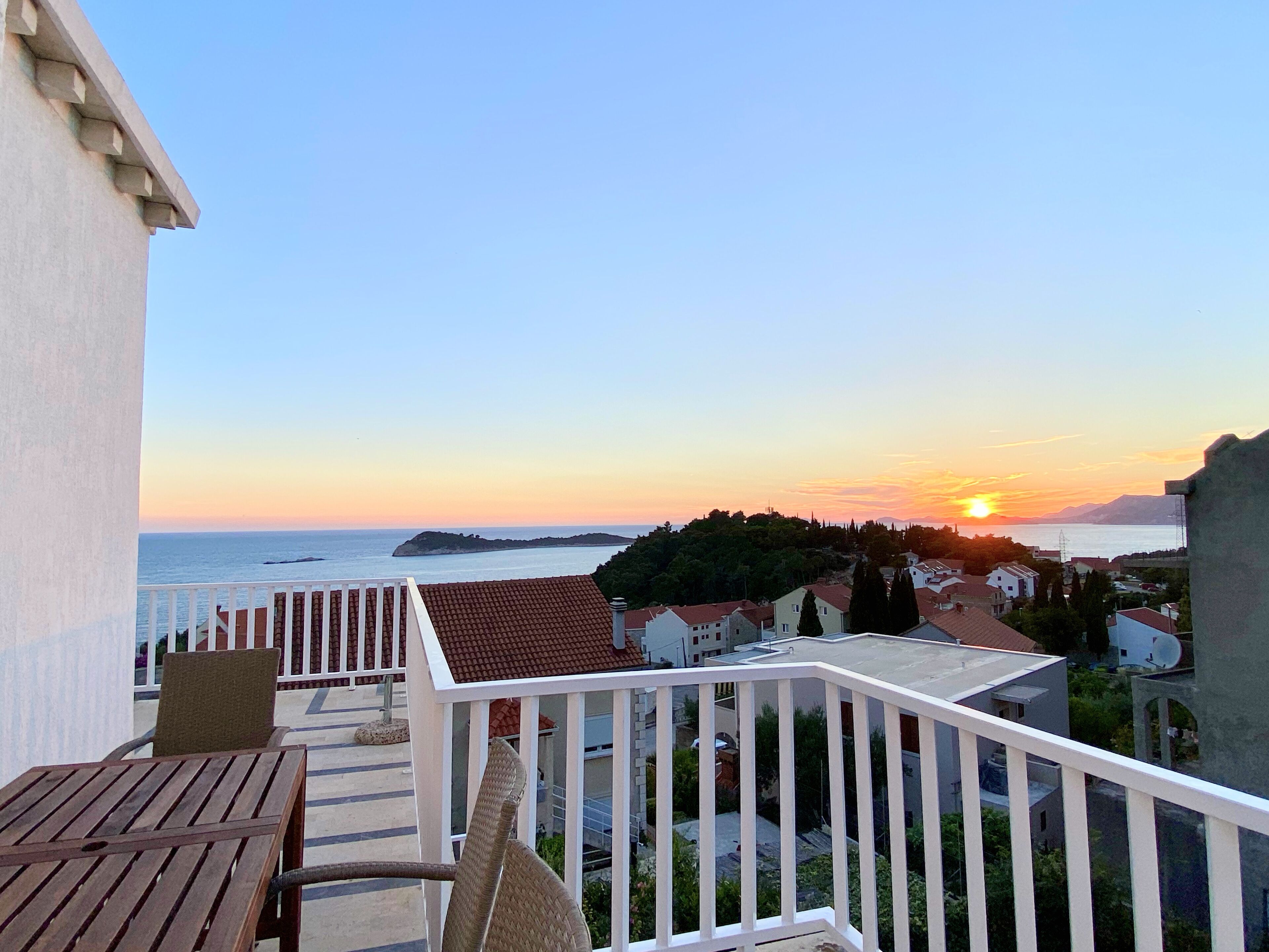 A balcony with a view of the sunset over the sea, a wooden table, and white railings.