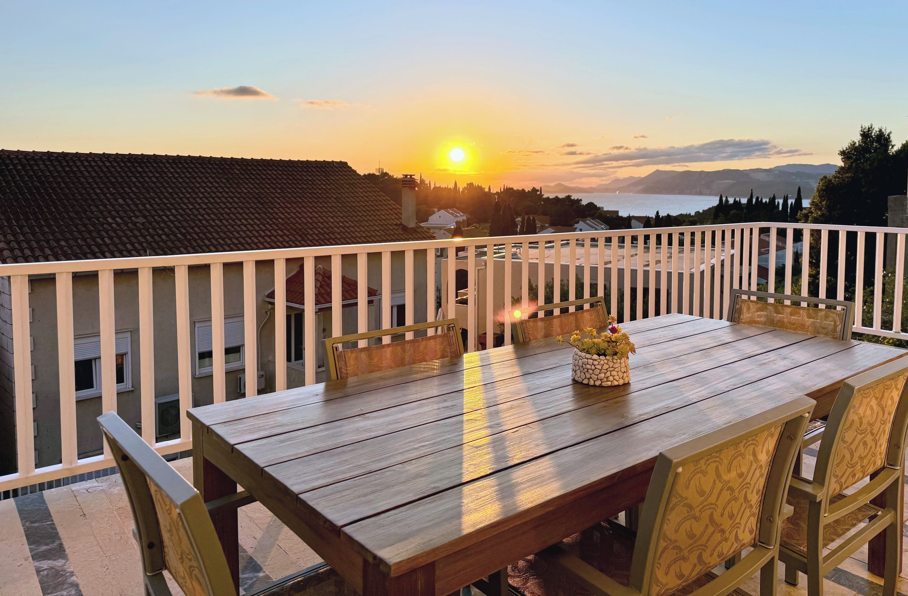 A wooden dining table with chairs on a balcony overlooking a sunset.
