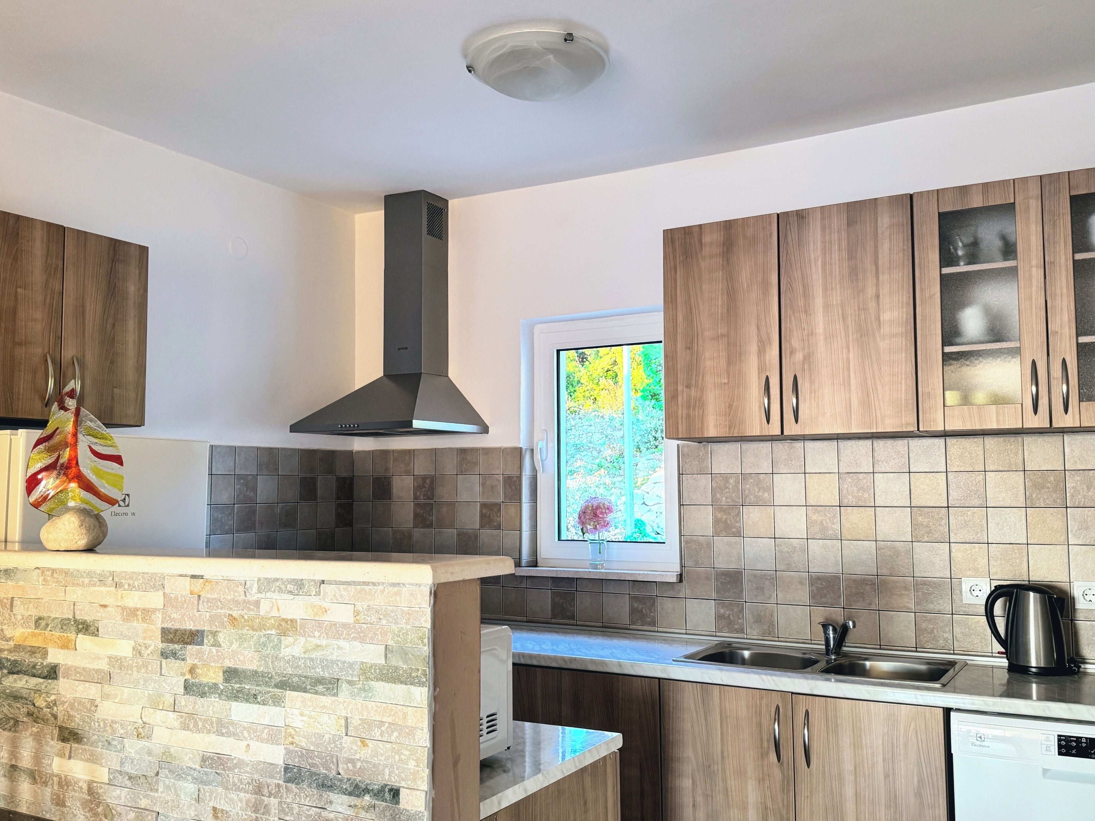 Een moderne keuken met een steensleuf achter het fornuis, houten keukenkastjes, een roestvrijstalen spoelbak en een inbouw vaatwasser.