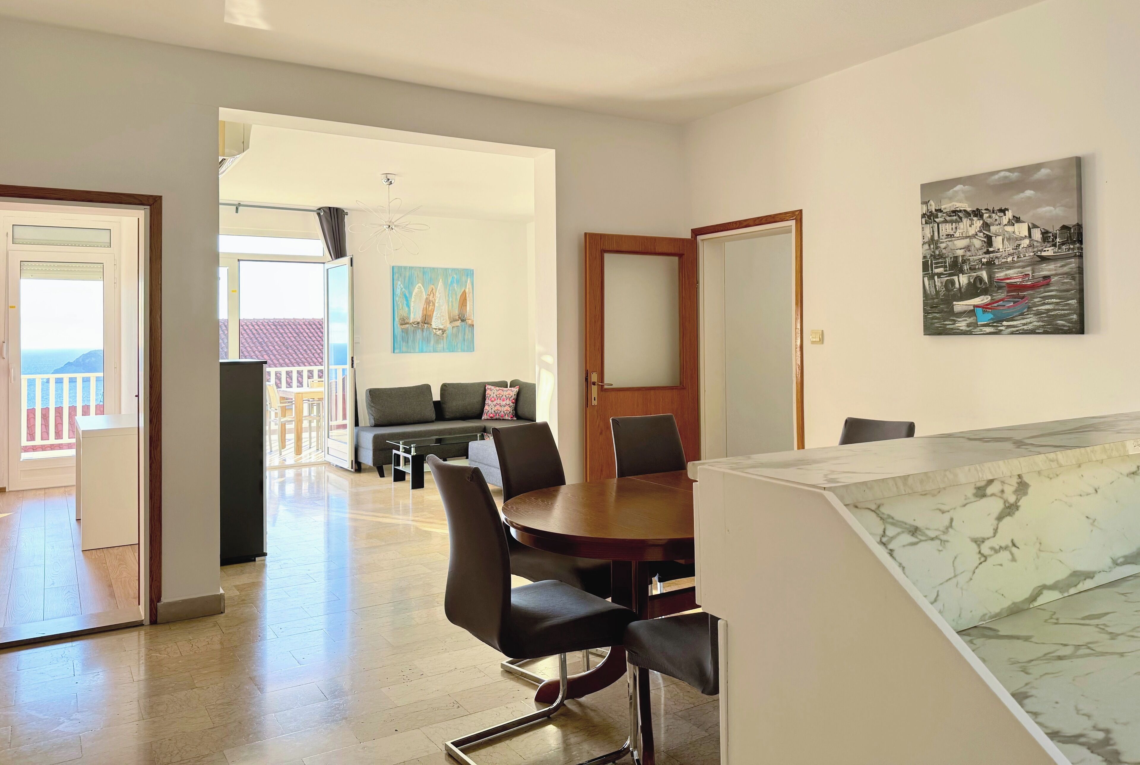 A dining area with a marble-top table and black chairs, a painting of boats on the wall, and a view of the balcony.