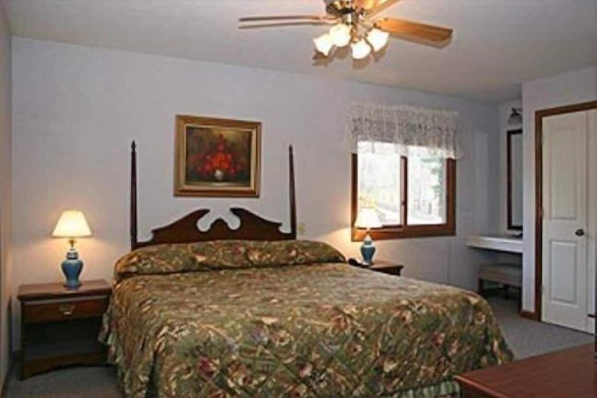 A bedroom with a bed, a nightstand, a lamp, a ceiling fan, a window with lace curtains, and a door.