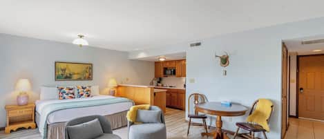 A hotel room with a bed, two armchairs, a dining table, a kitchen area, and a mounted deer head decoration.