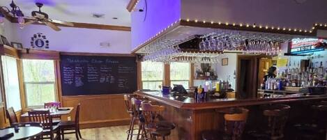 A bar area with wooden flooring, bar stools, a chalkboard menu, and a bar counter with glasses and bottles.