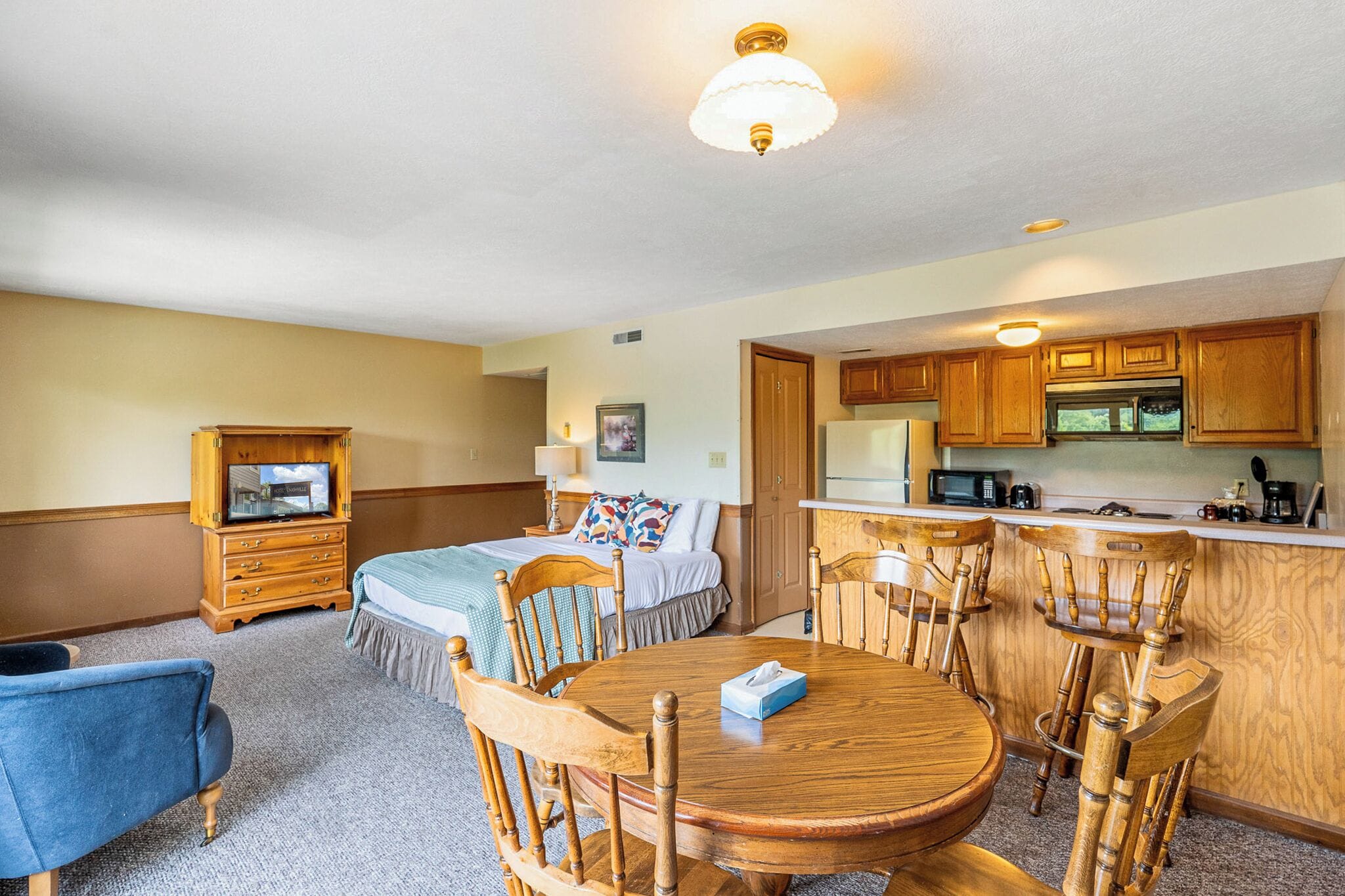 Habitación de hotel con cama, televisión, mesa de comedor con sillas, zona de cocina y un armario de madera.
