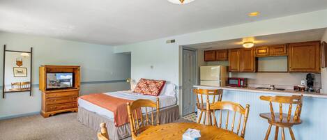 A room with a bed, a wooden dresser, a television, a dining table with chairs, and a kitchen area with wooden cabinets and a microwave.
