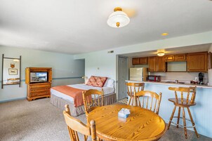 Una habitación con cama, un aparador de madera, televisor, una mesa de comedor con sillas y una zona de cocina con alacenas de madera y microondas.