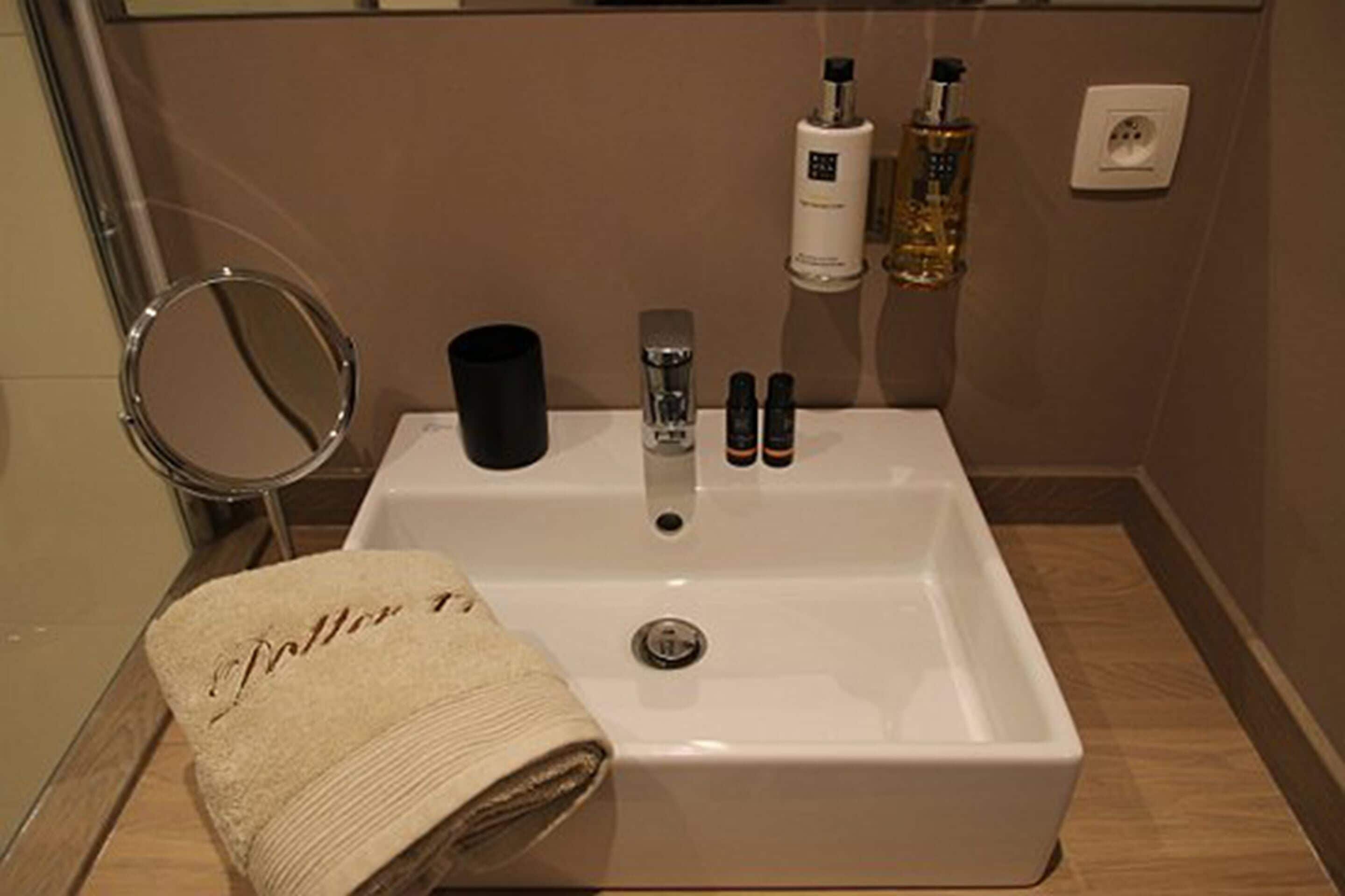 Un baño con un lavamanos blanco, un espejo redondo y una toalla con texto. Hay dos botellas y un recipiente negro sobre el lavamanos.