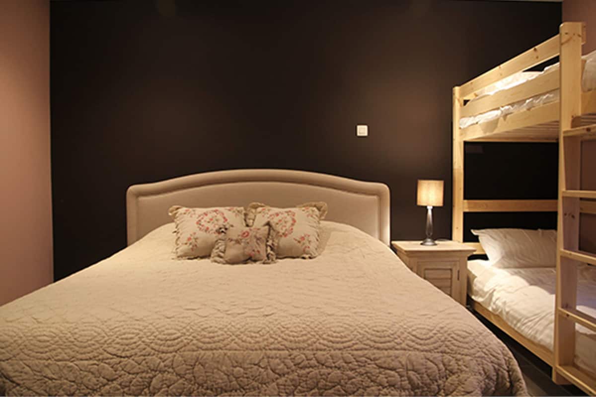 A bedroom with a large bed, a bedside table with a lamp, and a bunk bed to the right.