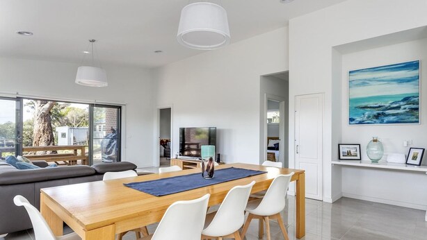 A modern living room with a wooden dining table, white chairs, a sofa, and a television. - Diamond Bay Beach House (SORRENTO)
