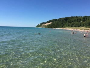 Una playa con agua cristalina, gente nadando y tomando el sol, y una colina boscosa al fondo.