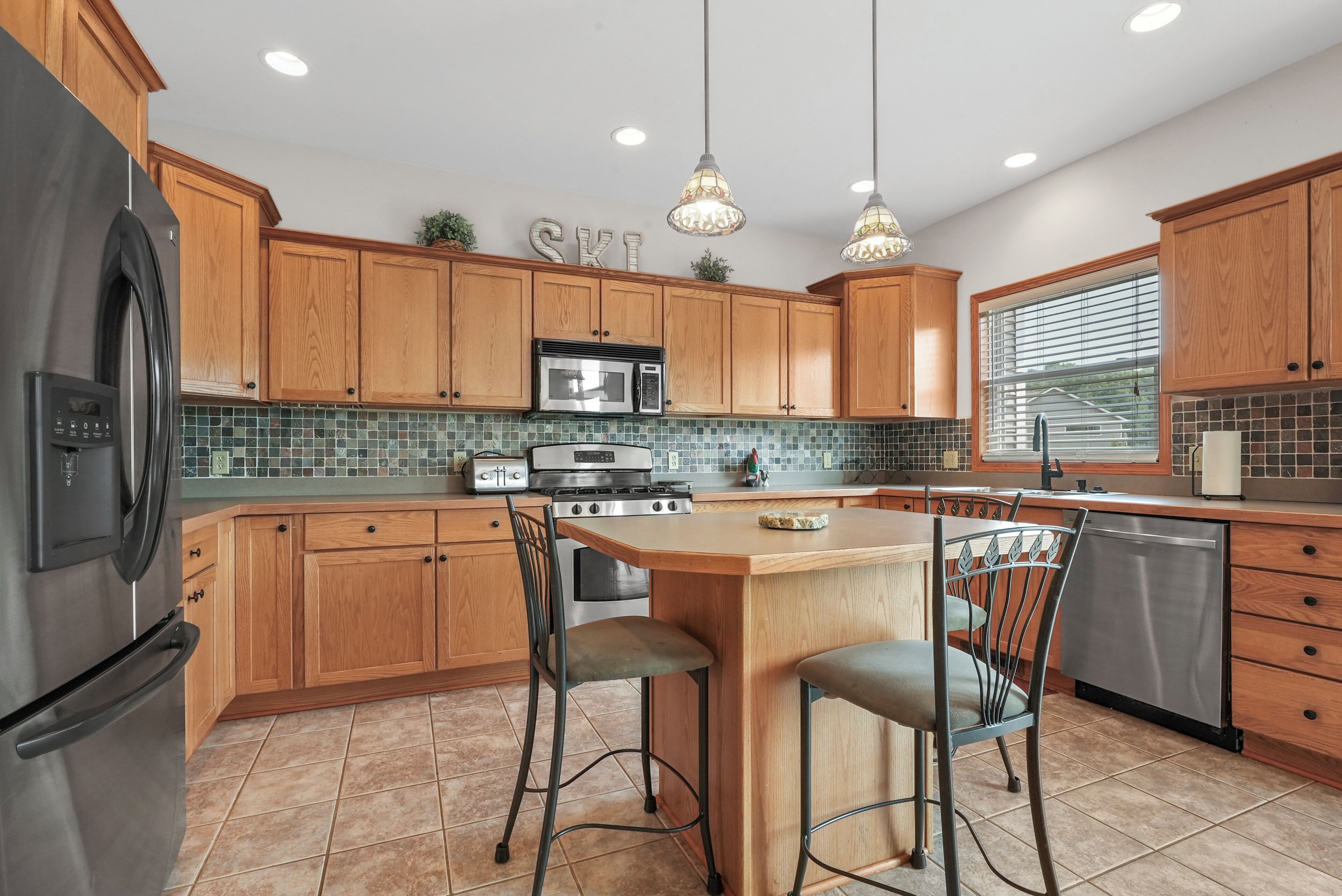 A modern kitchen with wooden cabinets, a stainless steel refrigerator, a microwave, and a dishwasher. There are two bar stools and a small island with a sink.