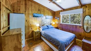 Une chambre à coucher aux murs lambrissés, comprenant un lit, deux lampes de chevet, une commode, une fenêtre donnant sur un beau paysage et un miroir.
