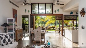 Amplia sala de estar con comedor, sofá, armario y vistas a un jardín tropical a través de puertas de vidrio grandes.