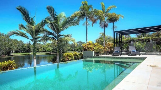 Una piscina cristalina con borde de azulejos, rodeada de palmeras y una pérgola con sillones de descanso.