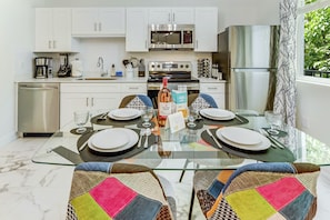 Una cocina moderna con armarios blancos, electrodomésticos de acero inoxidable y una zona de comedor con una mesa de vidrio y sillas coloridas.