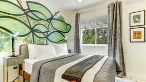 Une chambre à coucher avec une grande fresque murale abstraite, un lit recouvert d’une couette grise et blanche, une table de chevet avec une lampe et une fenêtre avec des rideaux.