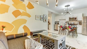Un salon moderne avec un canapé, une table basse et un tapis à motifs. On y trouve également un coin cuisine avec des meubles blancs, un réfrigérateur et une table à manger avec des chaises.