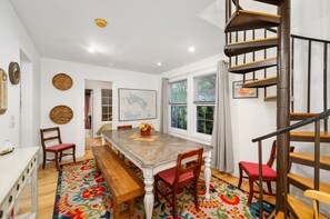 Un coin repas avec une table en bois, des chaises et un tapis coloré. Un escalier est visible sur la droite.