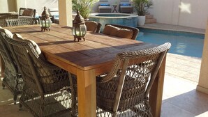 Patio esterno con tavolo in legno, sedie in vimini, una piscina e alcune piante in vaso.