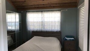 Ein Schlafzimmer mit Holzbalkendecke, einem Bett mit gemustertem Überwurf, einem Nachttisch mit Lampe und einem Fenster mit durchsichtigen Vorhängen.