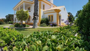 Une maison à deux étages, à la façade jaune et blanche, entourée de jardins bien entretenus et d’un palmier.