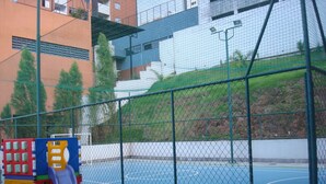 Un terrain de basket-ball entouré de clôtures, avec une aire de jeux comprenant un toboggan, situé à côté d’un bâtiment doté d’un toit végétalisé et d’un mur blanc.