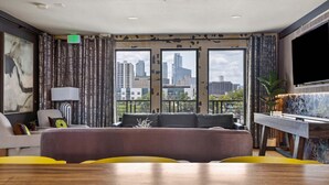 A modern hotel room with a large window offering a city view, a sofa, a dining table, and a TV. - (Unit 4) 6th Street Suite: 4BR/11Beds - Pool, Rooftop, Garden Bars, Walk to Everything! (Austin)