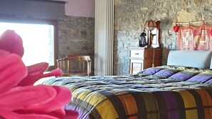 Une chambre avec un plafond en bois, un mur en pierre, un lit recouvert d’une courtepointe colorée, une table de chevet en bois, une chaise rouge et une fenêtre laissant entrer la lumière naturelle.