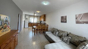 A living room with a sofa, a dining area with chairs, a kitchen in the background, and a painting on the wall.