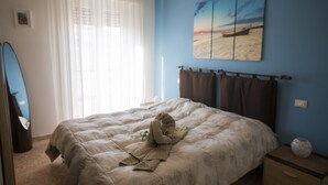 Une chambre à coucher avec un lit, une fenêtre ornée de rideaux légers, un tableau représentant une scène de plage et une petite table de chevet sur laquelle est posé un vase.