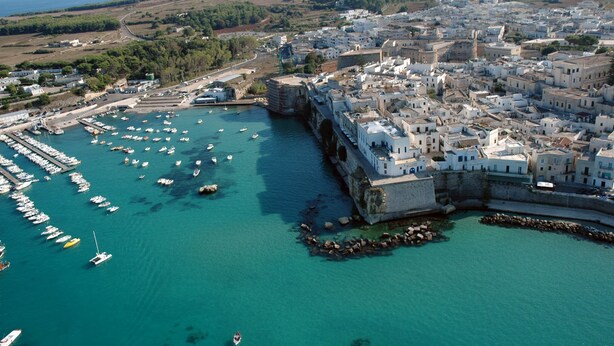 Une vue aérienne d’une ville côtière, avec un port rempli de bateaux, un mur fortifié et des bâtiments blancs surmontés de toits en dôme.