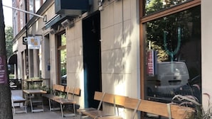 Un espace extérieur aménagé avec des bancs et des tables en bois, l’entrée d’un bâtiment et une voiture garée.